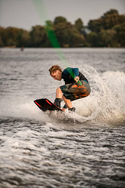 athletischer mann steht mit gebeugten knien auf wakeboard und reitet auf spritzender welle - knees bent stock-fotos und bilder