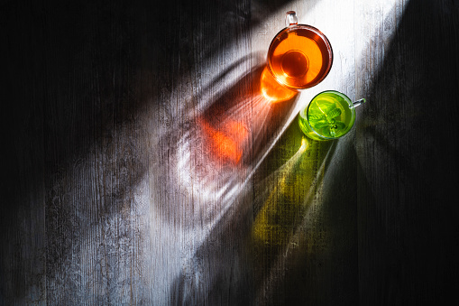 Red black tea cup and mint tea on gray wooden table and beam of light with moody dark surrounding.