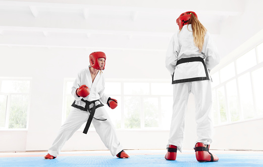 Young aggressive fighter standing ready to attack her opponent at training class. Fighter wearing in white kimono and red protective gloves and helmet with special shoes for fight boxing at gym.