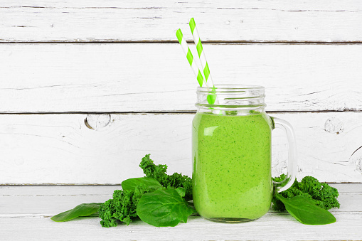 Green smoothie with kale and spinach in a mason jar glass. Side view with ingredients on a white wood background.