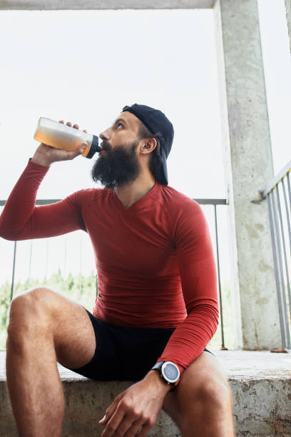 istock Side view of active bearded sportsman drinking water from glass bottle while having rest and sitting on stairs in bright day 1302131654
