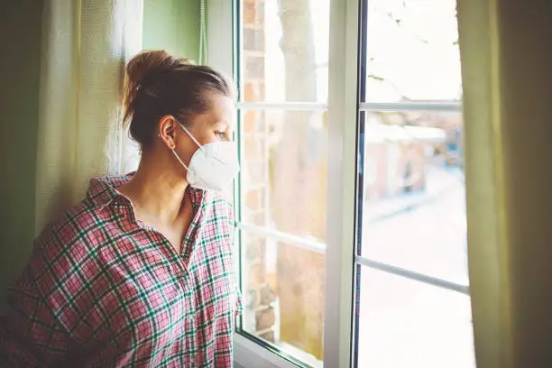 Photo of Pretty woman in quarantine with mask, looks out the window