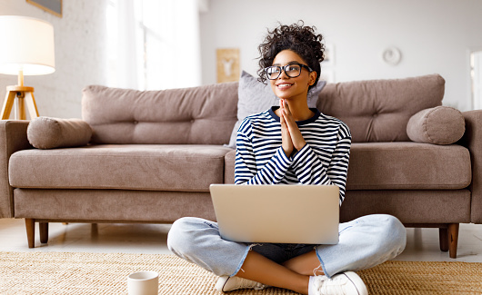 Pensive dreaming ethnic young female in glasses reading document and thinking while sitting cross legged near couch and working on remote project at home