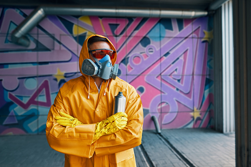 Graffiti painter in respirator mask standing near the wall with his paintings looking to camera. Street art concept