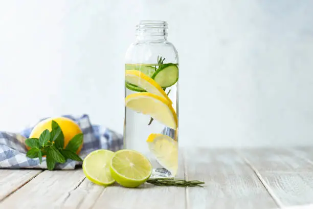 Bottle of infused water on white wood with a slice of lemon , cucumber and rosemary leaf in it.