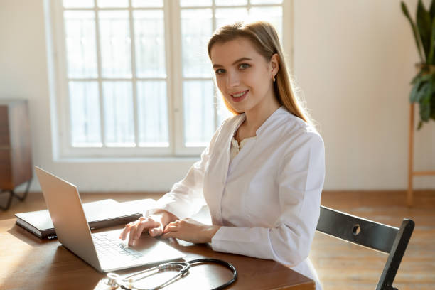 ritratto di piacevole giovane donna medico che usa laptop sul posto di lavoro - general practice foto e immagini stock