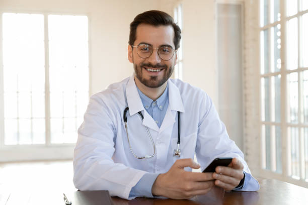 docteur confiant s’asseyant au bureau avec le téléphone capable d’aider - looking at camera smiling desk isolated photos et images de collection