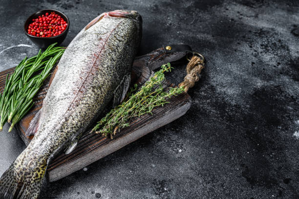 istock Raw Rainbow trout fish on a cutting board with herbs. Black background. Top view. Copy space 1302159369