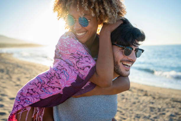 Multi ethnic Couples playing piggyback on the beach at sunset or sunrise. Multi ethnic Couples playing piggyback on the beach at sunset or sunrise. They are happy, smiling and laughing having fun with the ocean in the background. black couple sunset stock pictures, royalty-free photos & images