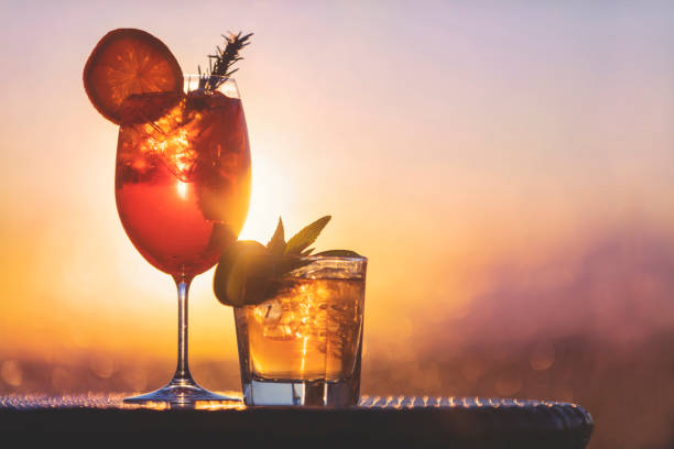 Two cocktails at sunset. One is a spritz with orange, the other is a mojito with mint and lime. The sun and sky is behind them