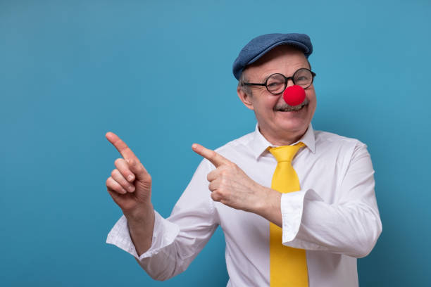 Senior cheerful man with red nose pointing with index finger aside. Portrait of a senior cheerful man with red nose pointing with index finger aside. Studio shot on blue wall. clowns nose stock pictures, royalty-free photos & images