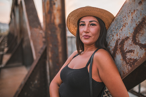 Young beautiful woman tourist in black dress on vacation wearing a hat standing on old rusty bridge