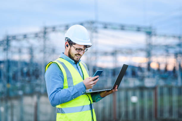 istock Electrical engineer working on a laptop and using a smart phone in front of Electricity power station stock photo 1302175851