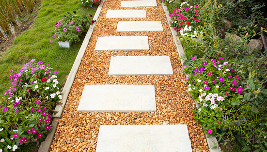 Selective focus shot of white stone path with yellow and brown gravel in colorful flower garden and green grass shows beautiful landscape of summer season. It is a gorgeous background of walkway.