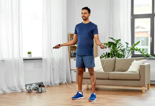 fitness, sport, exercising and healthy lifestyle concept - indian man skipping with jump rope at home