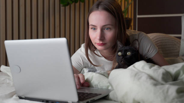 istock Attractive woman with a black cat uses a laptop while lying on the bed 1302214971