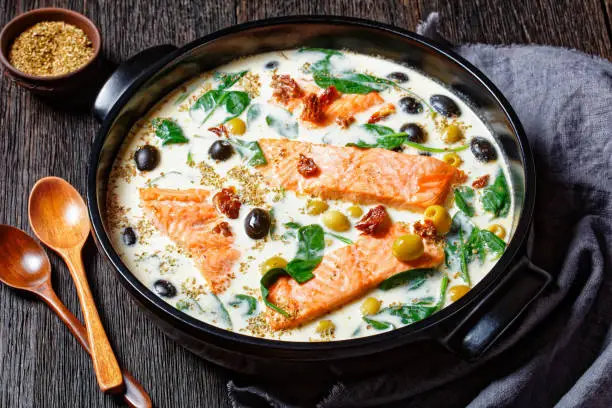 Tuscan salmon dish of pan-seared skin-on salmon fillet with cream sauce, garlic, leek, sun-dried tomatoes, olives, and spinach on a black  baking dish on a dark wooden table, top view, close-up