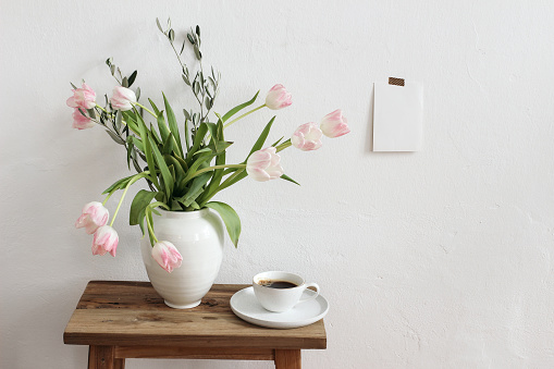 Easter, spring still life scene. Cup of coffee and floral bouquet in ceramic vase on wooden bench. Pink tulips flowers, olive tree branches on table,blank greeting card mockup taped on white wall.