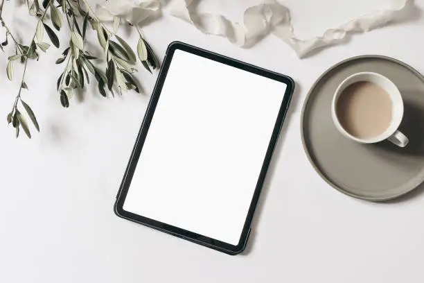 Electronic device mock-up. Closeup of tablet with empty screen. Cup of coffee, olive tree branches and ribbon isolated on white table background. Flat lay, top view, blogger design. Home office.