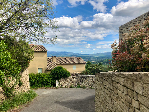 Horizontal photo of a quiet street away from the popular tourist centre of the charming hilltop town of Gordes in the south of France on a sunny day in Spring. Narrow roads meander past old hand-built stone walls, shrubs, fig trees, olive trees and cypress under a blue sky with white clouds.