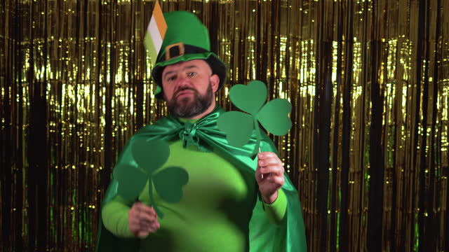 An elderly leprechaun celebrates St. Patrick's Day holding two clover leaves and dancing with them.