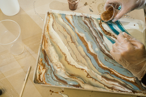 resin art, beautiful overflows of gold, beige and blue paint, golden liquid resin is poured from a plastic cup