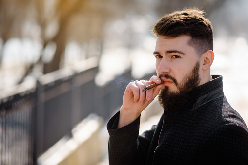 Close up portrait of bearded stylish young handsome man in black coat smoking  cigarette outside.