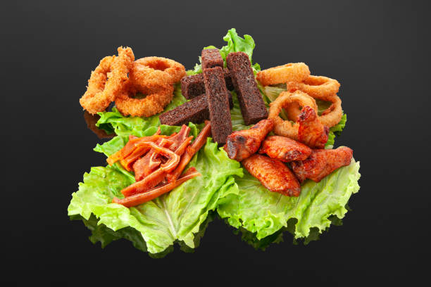 A set of snacks for beer. Grilled chicken wings, onion rings, squid rings, garlic croutons, dried pork ears. On a lettuce leaf. Decorated with greenery. Close-up. Dark mirror background. un juego de bocadillos para la cerveza. alitas de pollo a la parrilla, aros de cebolla, anillos de calamar, croutons de ajo, orejas de cerdo secas. sobre una hoja de lechuga. decorado con vegetación. primer plano. fondo de espejo oscuro.