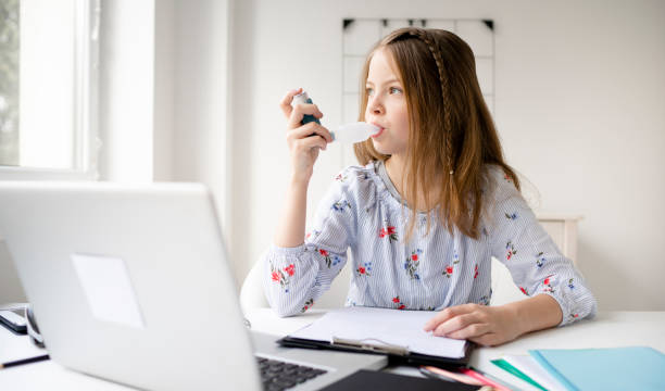 hübsche blonde mädchen sitzt am tisch arbeiten und inhalieren ein medikament - asthmatic child asthma inhaler inhaling stock-fotos und bilder