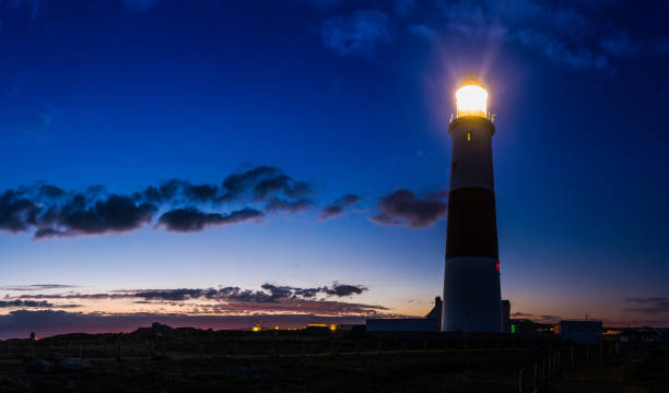 Lighthouse beaming out from ocean clifftop into blue dusk skies Lighthouse shining its beams across the cliffs of a rocky peninsula out to the dark ocean beyond as the warm light of sunset fades in the panoramic skies above, Portland Bill, Dorset. UK. bill of portland stock pictures, royalty-free photos & images