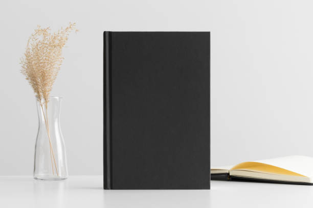 istock Black book mockup with a dried grass decoration and workspace accessories on a white table. 1302238836