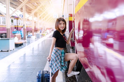 Travelers young woman getting on the train at train station. Beautiful woman with luggage smiling get in subway for traveling in holiday at platform looking someone. Travel and transportation concept.