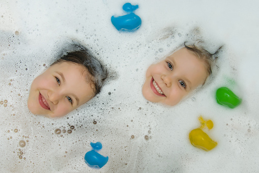 Two cute girls bathe together in a bath with ducks for bathing. The playful faces of children peek out of the foam, smiling cheerfully looking at the camera. View from above. Children's hygiene.