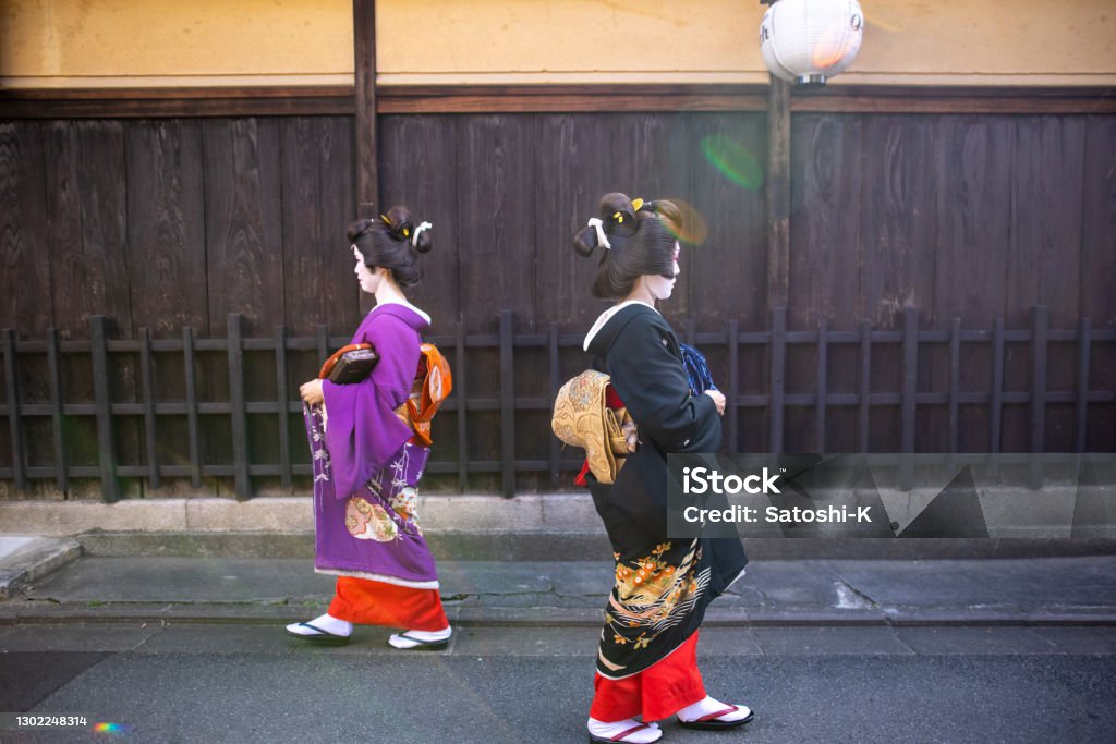 Two geisha women walking on traditional Japanese street in Gion district Japanese mid adult female friends visiting Kyoto and experiencing Geiko (Geisha) makeover. Wearing gorgeous kimono for Geisha with 'Oshiroi' special white face makeup. Geta Sandal Stock Photo