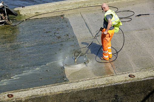 Plymouth England. The Cattewater. A man dressed in orange overalls cleaning seaweed off a concrete boat slipway using a water jet from a power washer drawing water from the sea. Cleaned area clearly defined. River Plym and dicks in background.