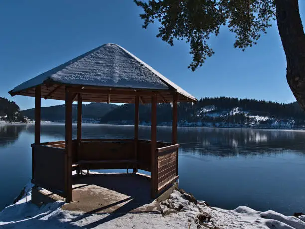 Photo of Snow-covered wooden pavillon on the shore of frozen Schluchsee lake, a popular tourist destination in Black Forest mountain range, Germany, in winter season.