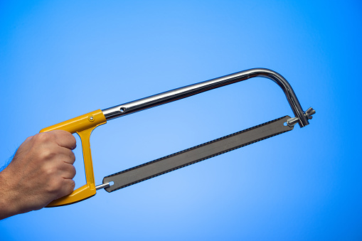 Caucasian male hand holding a hand saw with yellow plastic handle studio shot isolated on blue background.