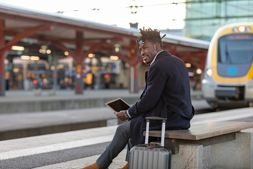 A man in smart clothing commuting to work. He is sitting on a bench in a train station and is typing on his laptop.