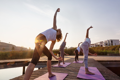 group of four women is practicing yoga on a jetty at, a yoga instructor is teaching them. The women are mixed age range, it is the time short after sunrise, the lake is Phoenixsee in Dortmund, Germany, they are doing the warrior pose, the photo is backlit