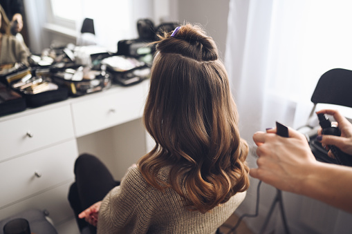 Hair stylist prepares woman makes curls hairstyle with curling iron. Long light brown natural hair