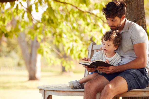 istock Father Sitting On Park Bench Under Tree With Son Reading Book Together 1302264813
