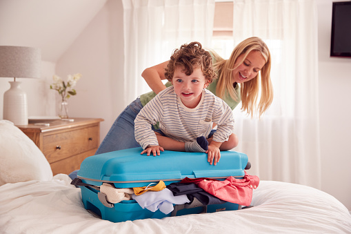 Mother And Son Packing For Vacation With Boy Standing On Full Suitcase To Close