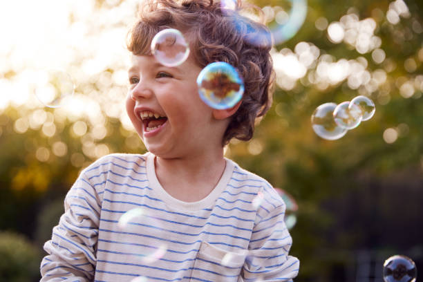 jovencito divirtiéndose en el jardín persiguiendo y reventando burbujas - niños fotografías e imágenes de stock