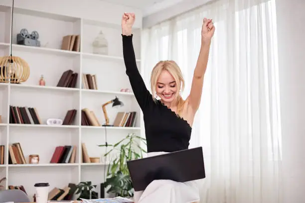 Young blond woman, wearing black top and white pants, sitting on office table, holding laptop, being happy, smiling, Office manager at her workplace. Female working on computer in company.