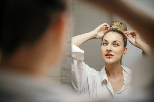 Beautiful young woman is doing her hair as part of her morning routine. She is looking in the mirror and checking her high bun. She is holding hair pin in her mouth, ready to use in and include in her hairstyle.
