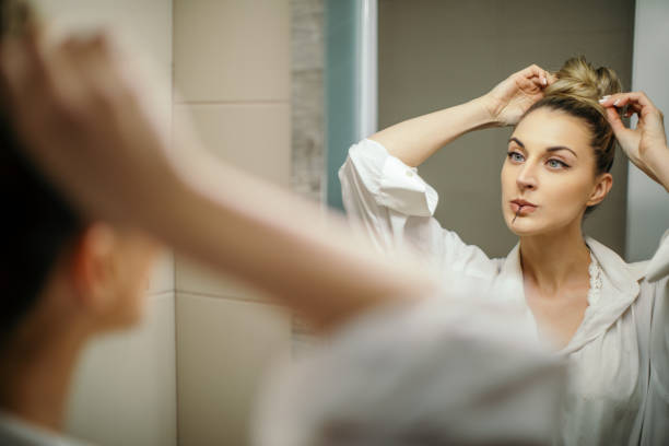 Beautiful young woman is doing her hair as part of her morning routine. She is looking in the mirror and checking her high bun. She is holding hair pin in her mouth, ready to use in and include in her hairstyle.