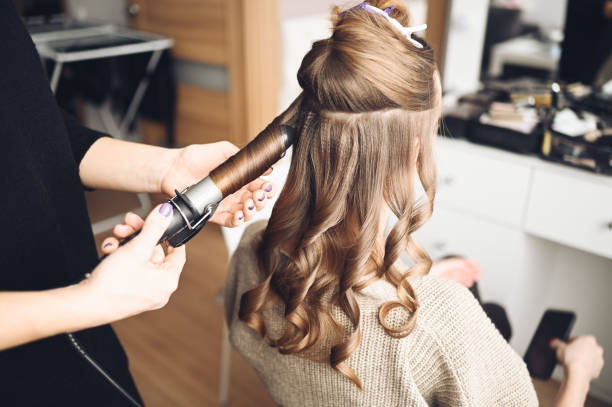 Hair stylist prepares beautiful young woman for event, makes curls hairstyle with a curling iron for client in beauty salon. Long beautiful light brown natural hair. stock photo