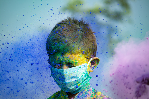 Portrait shot of young kid with face mask standing while colorful Holi powder throwing at him - concept of holi festival during coronavirus coivd-19 pandemic.