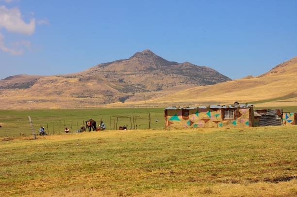 Kwa Sani Local Municipality, Mokhotlong, Lesotho - October 3,2008 : Four Lesotho Zulu shepherds with their  horse sitting on the ground next to their tin-roofed house with sheep pasture and mountains in the background