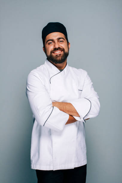 Mid adult chef in uniform on a greyish background Adult chef wearing his uniform. He is standing and looking at camera with a smile on his face looking friendly and proud chefs whites stock pictures, royalty-free photos & images